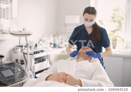 Man laying in cosmetologist cabinet and has a procedure for face skin.Female cosmetologist wearing blue medical costume. Woman wiping man's face with a cotton. 109404520