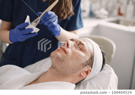 Man laying in cosmetologist cabinet and has a procedure for face skin. Unrecognized female cosmetologist wearing blue medical costume. Woman applying a mask on man's face. 109404524