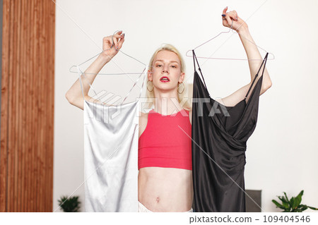 Man with make up on his face standing in a bedroom in his flat. Man wearing fashionable pink top, white trousers. Man choosing what to wear: black or white dress. 109404546
