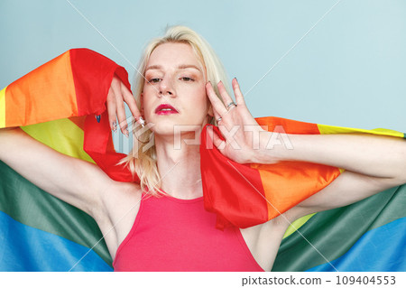 Young blonde man with make up on his face and rainbow flag posing for a photo. Man wearing fashionable pink top and has red lips. Man isolated on blue background. Young blonde man with make up on his face and rainbow flag posing for a photo. Man wearing fashionable pink top and has red lips. Man isolated on blue background. 109404553
