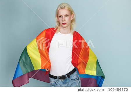 Young blonde man with make up on his face and rainbow flag posing for a photo. Man wearing fashionable white t-shirt and has red lips. Man isolated on blue background. 109404570