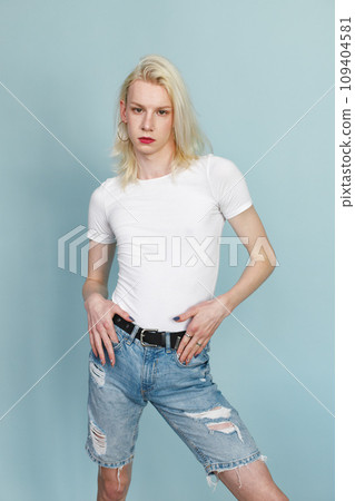 Young blonde man with make up on his face posing for a photo. Man wearing fashionable white t-shirt and has red lips. Man isolated on blue background. 109404581