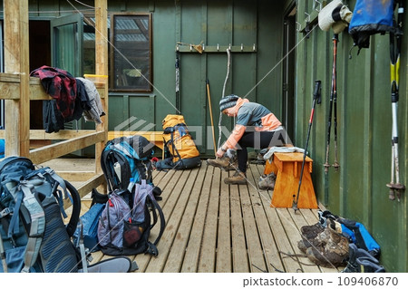 Woman putting on hiking shoes in front of a wooden cabin on a trail Woman putting on hiking shoes in front of a wooden cabin on a trail 109406870