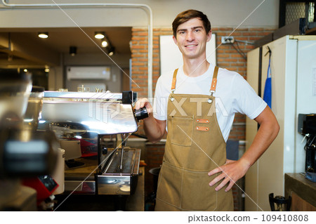 Professional young coffee barista standing inside the counter bar in the coffee shop and making arms crossed and looking at camera, handsome - good looking caucasian ethnic barista posing. Professional young coffee barista standing inside the counter bar in the coffee shop and making arms crossed and looking at camera, handsome - good looking caucasian ethnic barista posing. 109410808