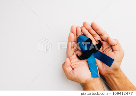 Supporting men living with cancer in November. A man's hands cradle a light blue ribbon with a mustache on a blue background, representing unity and strength. 109412477