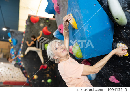 Determined old lady doing difficult wall climb in climbing gym 109413343