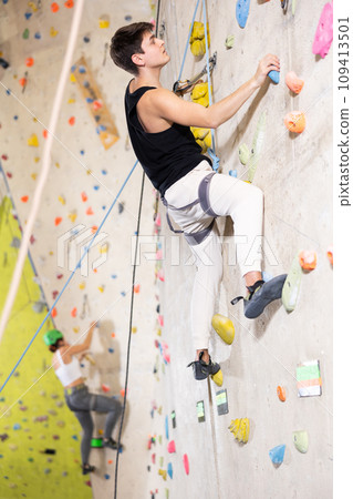young positive man, insured by trainer, moving up artificial climbing wall indoors young positive man, insured by trainer, moving up artificial climbing wall indoors 109413501