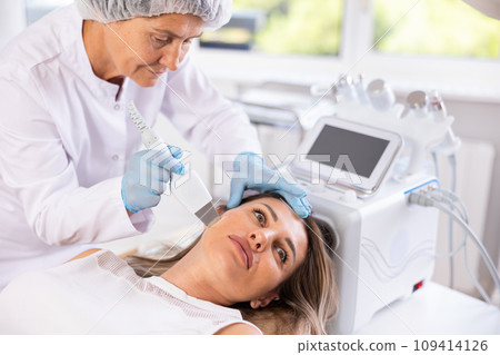 Young female client experiencing facial skin peeling with ultrasonic scrubber 109414126