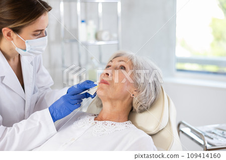 Eldery woman is getting a rejuvenating facial injections. She is lying calmly at clinic. Eldery woman is getting a rejuvenating facial injections. She is lying calmly at clinic. 109414160
