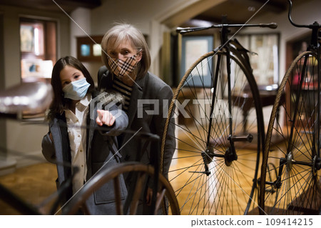 Elderly woman with preteen granddaughter in protective masks examining retro bicycles in museum 109414215