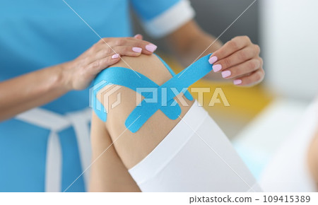 Traumatologist holds blue tape in his hand to fix patient's knee joint in clinic Traumatologist holds blue tape in his hand to fix patient's knee joint in clinic 109415389