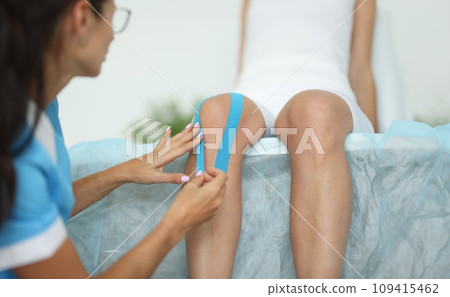 Physiotherapist sticking blue tape on patient's knee in clinic closeup 109415462