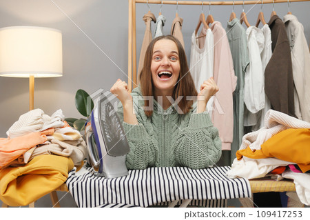 Extremely happy brown haired woman wearing knitted shirt ironing clothing while sitting in her wardrobe posing with clenched fists rejoicing looking at camera with amazed face Extremely happy brown haired woman wearing knitted shirt ironing clothing while sitting in her wardrobe posing with clenched fists rejoicing looking at camera with amazed face 109417253