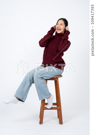 Young happy Asian woman is enjoying music on her headphones while sitting on a chair. 109417393