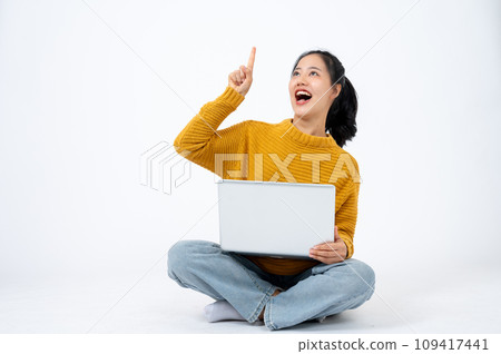 Beautiful woman sits on an isolated white background with her laptop and pointing her finger up. 109417441