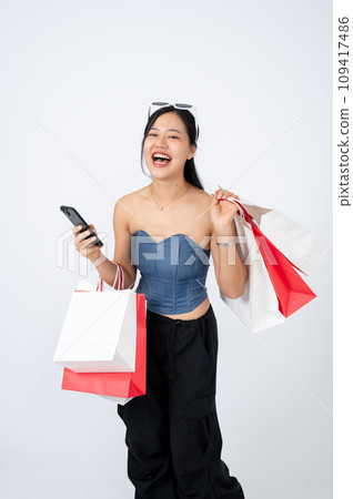 A cheerful and elegant Asian woman in trendy clothes is holding her smartphone and shopping bags. A cheerful and elegant Asian woman in trendy clothes is holding her smartphone and shopping bags. 109417486