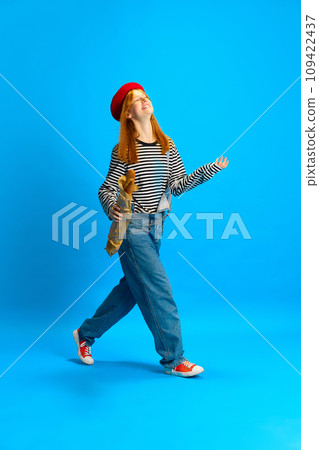 Happy cheerful woman, dressed retro fashion outfit in red beret walking with baguette against blue studio background. Happy cheerful woman, dressed retro fashion outfit in red beret walking with baguette against blue studio background. 109422437