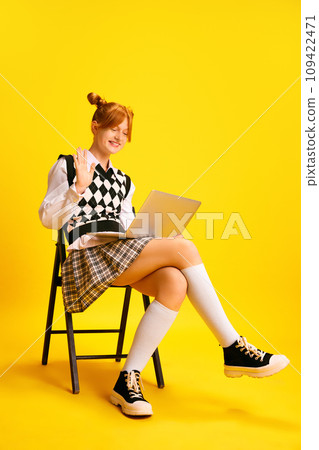 Happy cheerful girl sitting on chair and waving hello to laptop camera as she studies, holds conference online against yellow background. 109422471