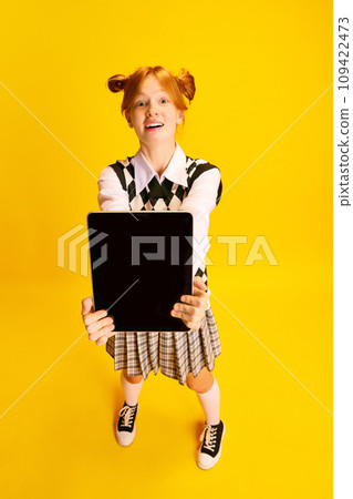Top view portrait of pleasantly shocked young attractive woman, student with red hair showing blank tablet screen against yellow background. Top view portrait of pleasantly shocked young attractive woman, student with red hair showing blank tablet screen against yellow background. 109422473