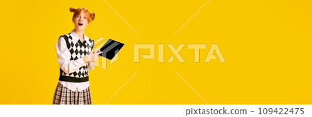 Banner. Adorable, pleasantly shocked young girl, student pointing on tablet with blank screen against yellow background with negative space for text. Banner. Adorable, pleasantly shocked young girl, student pointing on tablet with blank screen against yellow background with negative space for text. 109422475