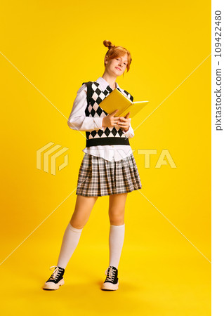 Faull-length portrait of beautiful red haired girl, dressed retro fashion outfit reading book against yellow background. Concept of education. Faull-length portrait of beautiful red haired girl, dressed retro fashion outfit reading book against yellow background. Concept of education. 109422480