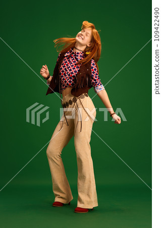 Full length portrait of cheerful young girl, teenager dressed like cowgirl and wind blowing her red hair while she dancing against green studio background. Full length portrait of cheerful young girl, teenager dressed like cowgirl and wind blowing her red hair while she dancing against green studio background. 109422490