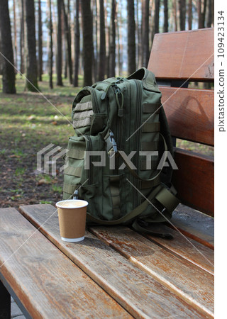 A cup of coffee next to a backpack leaning against the back of a wooden bench 109423134