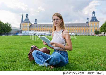 Young student girl sitting on grass, educational building background Young student girl sitting on grass, educational building background 109423629