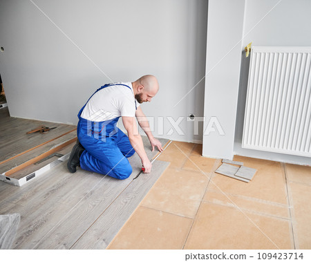 Male construction worker using tape measure while installing laminate flooring in apartment under renovation. Man in work overalls measuring distance from wall to laminate board. 109423714