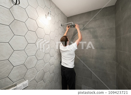 Back view of man standing by the wall with ceramic tile and installing high pressure bath showerhead at home. Male worker fixing bathroom showerhead while working on bathroom renovation. Back view of man standing by the wall with ceramic tile and installing high pressure bath showerhead at home. Male worker fixing bathroom showerhead while working on bathroom renovation. 109423721