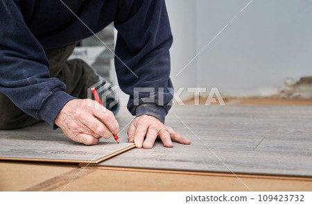 Close up of man construction worker with pencil in hand drawing mark on laminate wooden plank. Male worker preparing laminate boards for floor installation in apartment under renovation. Close up of man construction worker with pencil in hand drawing mark on laminate wooden plank. Male worker preparing laminate boards for floor installation in apartment under renovation. 109423732