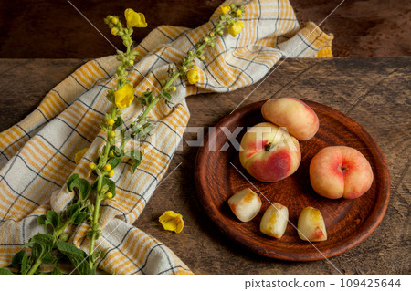 Clay plate with cut and whole saturn peaches or flat peaches on wooden background with yellow kitchen towel.. Clay plate with cut and whole saturn peaches or flat peaches on wooden background with yellow kitchen towel.. 109425644