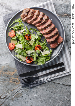 Tagliata di manzo Italian style beef steak served with arugula, cherry tomatoes and parmesan close-up in a plate. Vertical top view 109427062