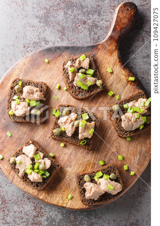 Rye toast with cod liver, green onions and capers close-up on a wooden board. Vertical top view 109427075