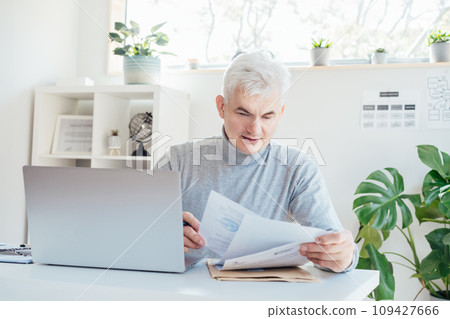 Middle aged businessman reading paper documents and using laptop while working at home office. Confident, experienced senior male professional. Small business entrepreneur manage business. paperwork Middle aged businessman reading paper documents and using laptop while working at home office. Confident, experienced senior male professional. Small business entrepreneur manage business. paperwork 109427666