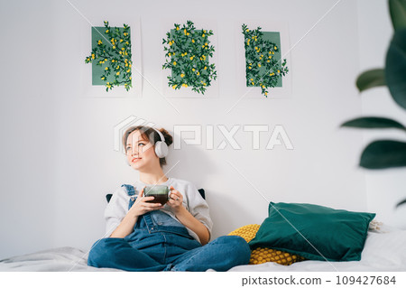 Smiling Woman listen music and enjoy cup of coffee or tea. Calm female spend free time at home enjoy favourite song with wireless modern headphones. Pastime weekend relax, no stress concept. 109427684