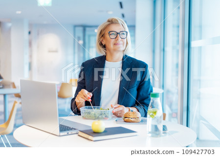 Smiling middle-aged business woman having healthy lunch at working place or business cafe, with laptop during her break. Balanced diet lunch box. Healthy eating habits and well-being. Selective focus 109427703