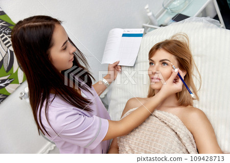 cosmetologist marking skin of caucasian woman with white pencil. facial marking for aesthetic procedure. cosmetologist marking skin of caucasian woman with white pencil. facial marking for aesthetic procedure. 109428713