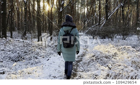 Young woman walking along path in sunny snowy forest. Girl with backpack going among trail at beautiful winter woodland. Unrecognizable lady enjoying stroll outdoor admiring beautiful scenic 109429654