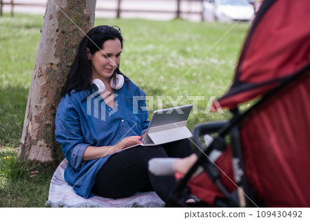 Positive woman with earphones typing on tablet keyboard while baby dreaming in stroller in green urban park mother studying online and walking infant child 109430492