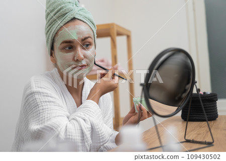 Facial Bliss: She, in a bathrobe, applies a clay mask with a brush, gazing at the mirror, indulging in her organic facial treatment at home.  109430502