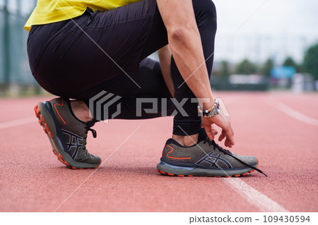 A closeup of an athlete taking a break from training, squatting on the rubberized running track, tying his sneaker laces at the urban outdoor stadium A closeup of an athlete taking a break from training, squatting on the rubberized running track, tying his sneaker laces at the urban outdoor stadium 109430594