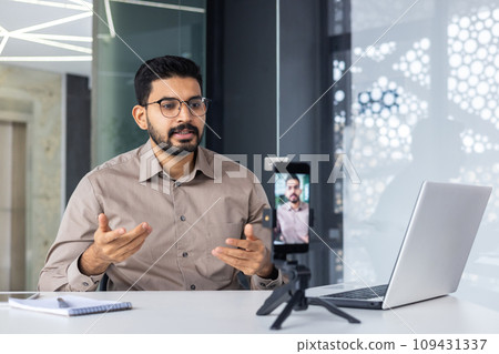 Successful business trainer teacher recording online training video course, man inside office at workplace using phone and tripod, sitting at desk with laptop. 109431337