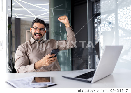 Successful businessman winner inside office at workplace, man smiling and looking at camera received online win notification win, employee at table with laptop celebrating triumph. Successful businessman winner inside office at workplace, man smiling and looking at camera received online win notification win, employee at table with laptop celebrating triumph. 109431347