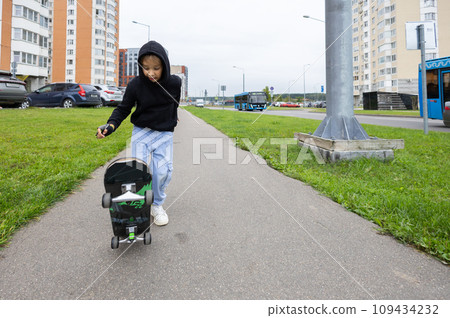 A beautiful child rides a skateboard along the city sidewalk. A child in a hood on a skateboard walks in the city during the day. Childhood of children in the city. City life. 109434232