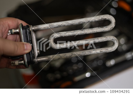 A hand holds a heating element with limescale deposits in front of a washing machine. Damage to the washing machine due to the accumulation of limescale on the heating element. 109434291