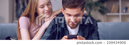 Lazy weekend. Young beautiful couple in casual wear looking at smartphone while relaxing together on the sofa at home in the living room 109434908