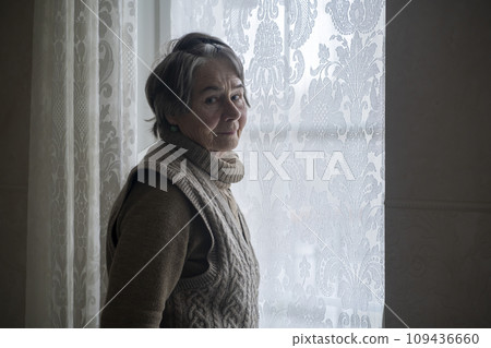 An elderly beautiful retired woman looks in the window with lace curtains. 109436660