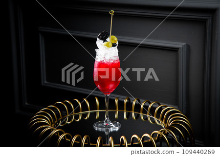 red cocktail in a transparent glass with round ice, whipped cream and black berry on top, side view 109440269