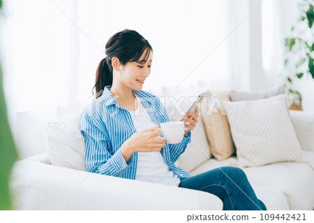 一個女人坐在沙發上，一邊喝咖啡一邊看著她的智能手機 109442421